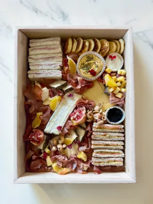 Elegant wooden box platter containing gourmet selections of smoked salmon sandwiches, ham with parmesan, assorted premium cheeses including truffle brie, homemade blinis with caviar, butternut houmous, charcuterie, and garnished with fresh fruits and nuts. Sauge Marbella's Gourmet Deluxe Platter - Riviera.