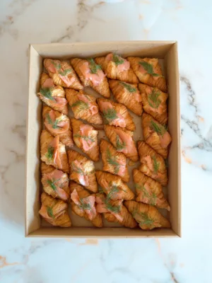 Box of 24 golden mini croissants topped with pink smoked salmon slices, fresh green dill sprigs, and creamy gravlax sauce arranged in neat rows. Perfect for catering events in Marbella.